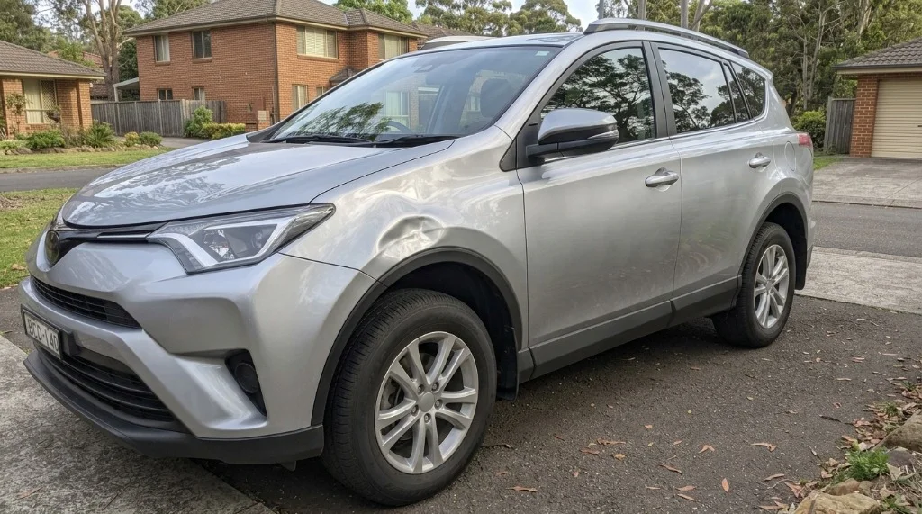 A silver Toyota RAV4 front fender photographed at a 3/4 angle catching sunlight across the dent depth.